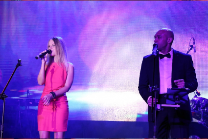 Danydeee singing on stage with a musician in a tuxedo under purple lights